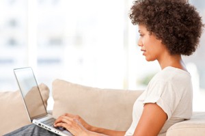 Beautiful young woman sits on her sofa while typing on a laptop. Horizontal shot.